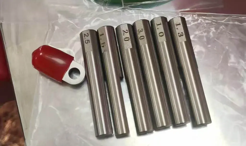 Set of six precision metal wire or tube samples with labeled diameters ranging from 1.0mm to 2.5mm, displayed on a plastic sheet with a red tool