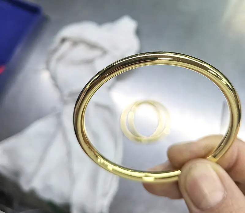 A hand holding a polished gold bangle bracelet up to the light, showing its highly reflective mirror-like finish and smooth circular form with a blurred workshop background.